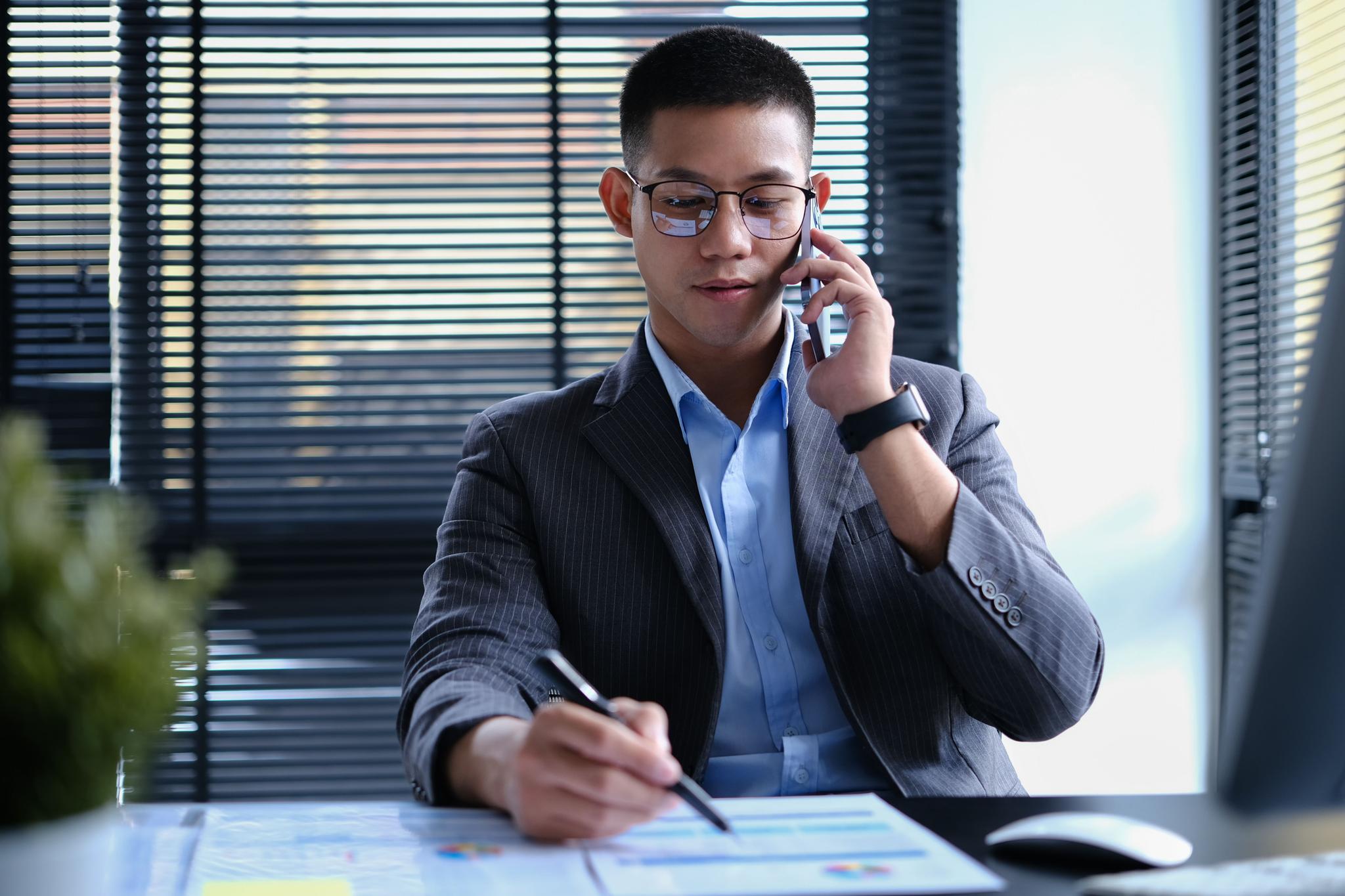 Asian male investor talking on mobile phone, negotiating with business partner modern workplace.